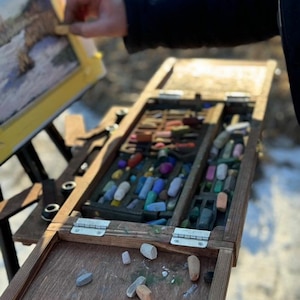 Könnte beinhalten: Ein Holz-Staffelei mit einer Palette voller Pastellkreiden. Die Palette ist geöffnet und einige Pastellkreiden liegen auf der Oberfläche verstreut. Die Staffelei steht im Freien auf einer schneebedeckten Fläche.