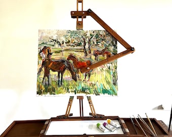 Puede incluir: Un caballete de madera con una pintura de caballos en un campo. El caballete tiene una bandeja con tubos de pintura y pinceles. La pintura está en un lienzo y está enmarcada.