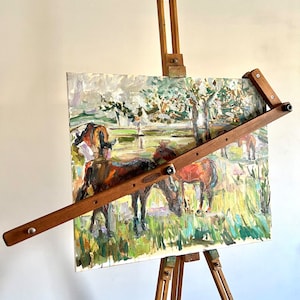 May include: A wooden easel holds a rectangular canvas featuring an oil painting of brown horses grazing in a grassy field with trees. A diagonal wooden mahl stick is attached across the front of the canvas, secured by metal hardware.