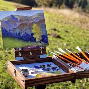 Può includere: Cavalletto da campo in legno con un dipinto di paesaggio in corso. Il dipinto raffigura montagne, alberi e un campo. Pennelli e una tavolozza con vernice sono visibili nella scatola di legno aperta. La scena è all'aperto.