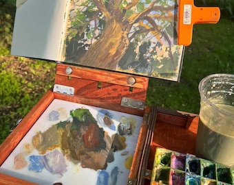 Può includere: Una scatola da pittura in legno che sostiene un album con il dipinto di un albero, fissato da una pinza arancione brillante. La scatola presenta una tavolozza bianca con macchie di colore, un vassoio per colori a griglia e un bicchiere d'acqua.