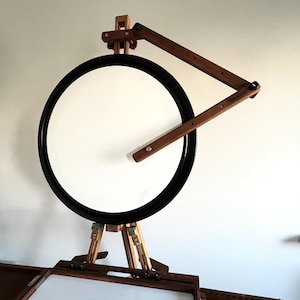 A wooden artist's easel with a circular rotating canvas. The easel is dark brown wood with light brown accents. The canvas is white and framed in black.  The easel features an adjustable arm for angled viewing.  It's a portable studio easel, perfect for plein air painting or studio use. The design is compact and functional.