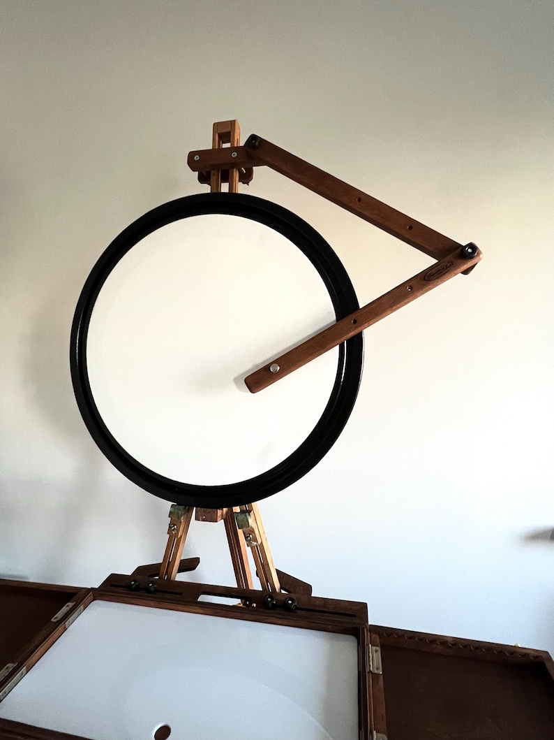 A wooden artist's easel with a circular rotating canvas. The easel is dark brown wood with light brown accents. The canvas is white and framed in black.  The easel features an adjustable arm for angled viewing.  It's a portable studio easel, perfect for plein air painting or studio use. The design is compact and functional.