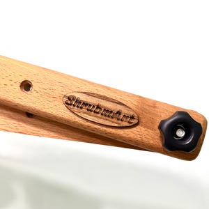 a light brown wooden Mahlstick with a black knob at the end.  The armrest features a dark brown oval logo with the text 'ShrubmArt' engraved into it.  The wood grain is visible, and there are two small holes visible on the armrest. The overall design suggests a piece of furniture or a component of a larger wooden structure.