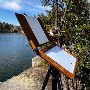 Könnte beinhalten: Ein braunes Holzmalstaffelei mit weißer Leinwand, im Freien in der Nähe eines Gewässers aufgestellt. Die Staffelei hat schwarze Klammern und ist auf einem schwarzen Stativ montiert. Der Hintergrund umfasst Bäume und einen blauen Himmel.