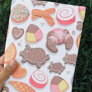 May include: A white pouch with a repeating pattern of various baked goods and candies, including cookies, croissants, and candies. The design features a variety of colors, including pink, brown, and yellow. The pouch is held against a background of green grass.