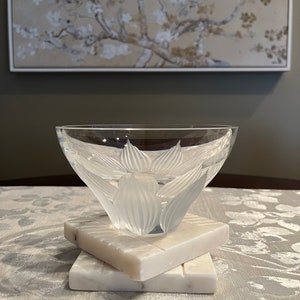 May include: A clear glass bowl with a frosted floral design sits on two white marble coasters. The bowl is on a white tablecloth with a floral pattern.