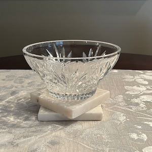 May include: A clear, cut-glass bowl with a starburst pattern, resting on two square, white marble coasters. The bowl is centered on a patterned, light-colored tablecloth.