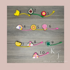 May include: A collection of colorful silicone charms with various designs, including a banana, avocado, flower, rainbow, orange slice, hot air balloon, lemon slice, rainbow, four-leaf clover, and rainbow. Each charm is attached to a flexible cord.