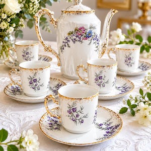 Peut inclure: Service à thé en porcelaine blanche avec bordure dorée, comprenant une théière, des tasses et des soucoupes. L'ensemble est orné de motifs floraux violets. La théière a une anse et un bec courbés. Les tasses et les soucoupes sont disposées sur une nappe blanche.