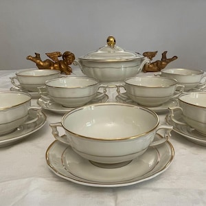 Peut inclure: Service à soupe ancien en porcelaine crème avec bordure dorée. L'ensemble comprend une soupière couverte, huit bols à soupe et des soucoupes. Deux figurines de chérubins dorés sont de chaque côté de la soupière.