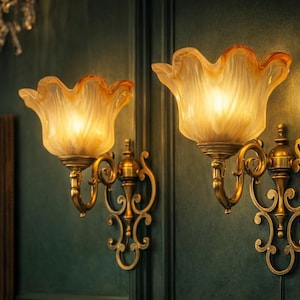 Könnte beinhalten: Zwei verzierte Wandleuchten mit bernsteinfarbenen Glasschirmen und Messing-Metallbeschlägen. Sie sind an einer dunkelgrünen Wand angebracht. Ein Kronleuchter und ein gerahmter Spiegel sind im Hintergrund zu sehen. Die Leuchten sind beleuchtet und strahlen warmes Licht aus.