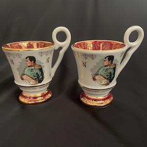 Peut inclure: Deux tasses à thé en porcelaine blanche avec un intérieur et une base rouges et dorés. Chaque tasse présente un portrait de Napoléon Bonaparte et la lettre "N". Les anses sont de forme circulaire.