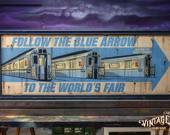 May include: Vintage-style sign with a blue arrow and text "FOLLOW THE BLUE ARROW TO THE WORLD'S FAIR." Features illustrations of subway cars. The sign is framed in black and has a distressed, aged appearance.
