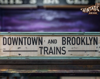 May include: A vintage-style metal sign with a distressed white background and black lettering. The sign reads "Downtown and Brooklyn Trains" with arrows pointing in opposite directions.