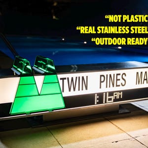 Puede incluir: Un letrero rectangular para "TWIN PINES MALL" con un reloj digital que muestra la 1:16 AM. El letrero tiene acentos geométricos verdes y el texto "NOT PLASTIC", "REAL STAINLESS STEEL" y "OUTDOOR READY".