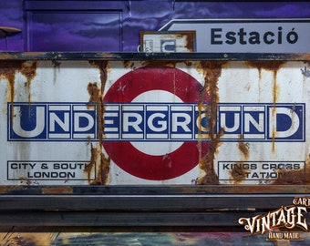 May include: A vintage, rusty metal sign with the words "UNDERGROUND" in blue and white letters. The sign also features a red circle with a white inner circle, and the text "CITY & SOUTH LONDON" and "KINGS CROSS TATION".