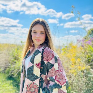 May include: A patchwork jacket with a geometric design. The jacket features a mix of pink, teal, and burgundy fabrics, with floral and paisley patterns. The jacket has a black floral trim and a relaxed fit.