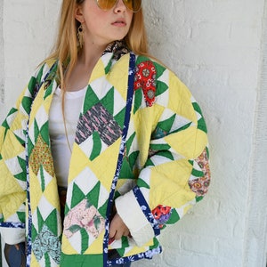 May include: A woman wearing a yellow, green and white patchwork quilted jacket with a blue trim. The jacket features a variety of floral and geometric patterns. She is also wearing a white t-shirt, blue jeans and yellow sunglasses.