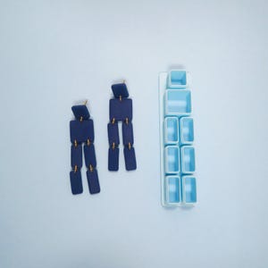 May include: A pair of navy blue geometric drop earrings with gold-coloured connectors. The earrings feature square and rectangular shapes. A light blue organiser with small, square compartments is also visible.