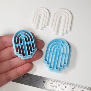 May include: Two sets of rainbow-shaped cookie cutters. One set is light blue and the other is white with black speckles. The cutters are shown on a white surface with a ruler for scale.