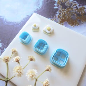 Op de afbeelding: Een verzameling blauwe plastic koekjessnijders in de vorm van toast en gebakken eieren. De snijders zijn gerangschikt op een wit oppervlak met decoratieve bloemen.
