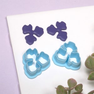 May include: Two purple plastic flower-shaped cookie cutters and two light blue plastic flower-shaped cookie cutters on a white surface.