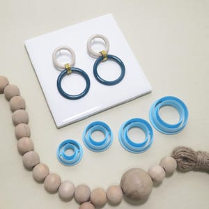 May include: A pair of large, dark teal and beige hoop earrings with gold-colored accents. Several light blue circle-shaped polymer clay cutters are also visible. A strand of wooden beads and a jute twine bundle are in the foreground.