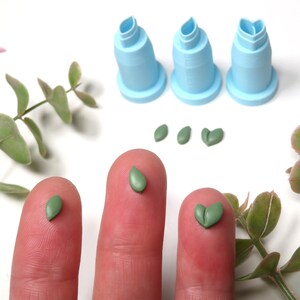 May include: Three small green leaf shapes made of polymer clay, two are single leaves and one is a heart-shaped double leaf. The leaves are on a fingertip, and there are three blue plastic leaf-shaped tools in the background.