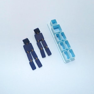May include: Navy blue geometric dangle earrings with gold-colored accents. The earrings are composed of square and rectangular shapes. A light blue plastic earring cutter is also in the image, with various square and rectangular shapes.