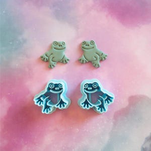 Peut inclure: Quatre objets en forme de grenouille. Deux sont vert clair uni et deux sont bleu clair avec un intérieur bleu plus foncé. Tous ont des visages souriants et des pieds détaillés. Le fond est rose et blanc doux.