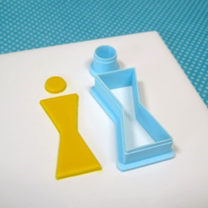 May include: A light blue plastic cookie cutter shaped like a person with a round head and a bow tie. A yellow fondant cutout of the same shape is next to the cutter.
