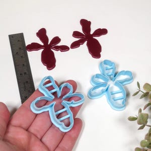 May include: An overhead shot shows two maroon flower-shaped clay pieces and two light blue flower-shaped cookie cutters. A metal ruler is next to the clay pieces. The cookie cutters are held in a hand. A small plant is in the background.