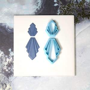 May include: Two Art Deco style earring cutters and a pair of earrings. One pair is a solid blue, the other is a light blue cutter. The earrings and cutters are on a white surface with a marbled background.