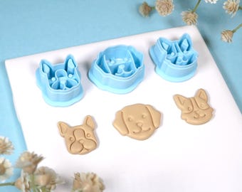 Puede incluir: Un conjunto de cortadores de galletas y galletas con forma de perro. Los cortadores son de plástico azul claro y las galletas son de color beige. Los cortadores tienen la forma de un bulldog francés, un labrador y un corgi.