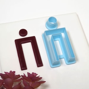 May include: Two cookie cutters, one in light blue and one in burgundy. The light blue cutter is shaped like the letter "H" with a small circle on top. The burgundy cutter is shaped like the letter "n" with a small circle on top.
