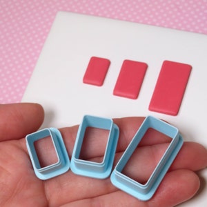 May include: Three pink rectangular shapes and three light blue rectangular cookie cutters in varying sizes are displayed. The cutters are held in a hand, with the pink shapes resting on a white surface. The background is a pink polka dot pattern.