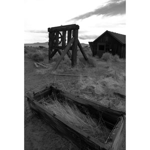 Op de afbeelding: Een zwart-witfoto van een houten constructie met een groot wiel, een klein houten gebouw en een houten trog op de voorgrond. De scène speelt zich af in een veld met hoog gras.