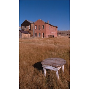 May include: A weathered wooden table with white paint sits in a field of dry grass in front of an abandoned brick building.