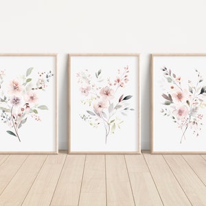 May include: Set of three watercolor prints featuring delicate pink and gray floral bouquets with green leaves. The prints are framed in natural wood frames and are perfect for adding a touch of nature to any room.