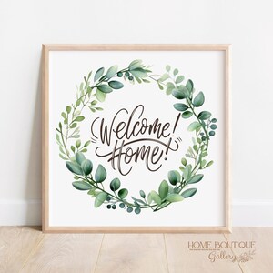 May include: Square framed print with the words "Welcome! Home!" in elegant script, surrounded by a watercolor wreath of green leaves. The print is displayed on a light wood surface.