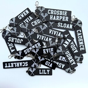 May include: A collection of black rectangular silicone keychains featuring white text names and a white graphic of two crossed lacrosse sticks. Each keychain includes a silver metal lobster clasp attached to a circular ring at the top end.