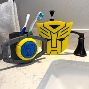 May include: A yellow and black soap dispenser and toothbrush holder set with a blue and yellow accent, featuring a robot face design. Two toothbrushes are in a gray toothbrush holder. The set is on a white countertop.