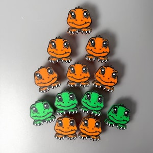 May include: A collection of orange and green lizard-shaped embellishments with black outlines. The lizards have smiling faces and clawed feet. The embellishments are arranged in a triangular pattern against a gray background.