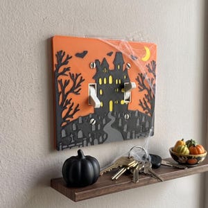 May include: A rectangular orange light switch cover plate featuring a black haunted house silhouette, bare trees, tombstones, bats, and a yellow crescent moon. The cover has two white toggle switches and is decorated with thin white faux spiderwebs.