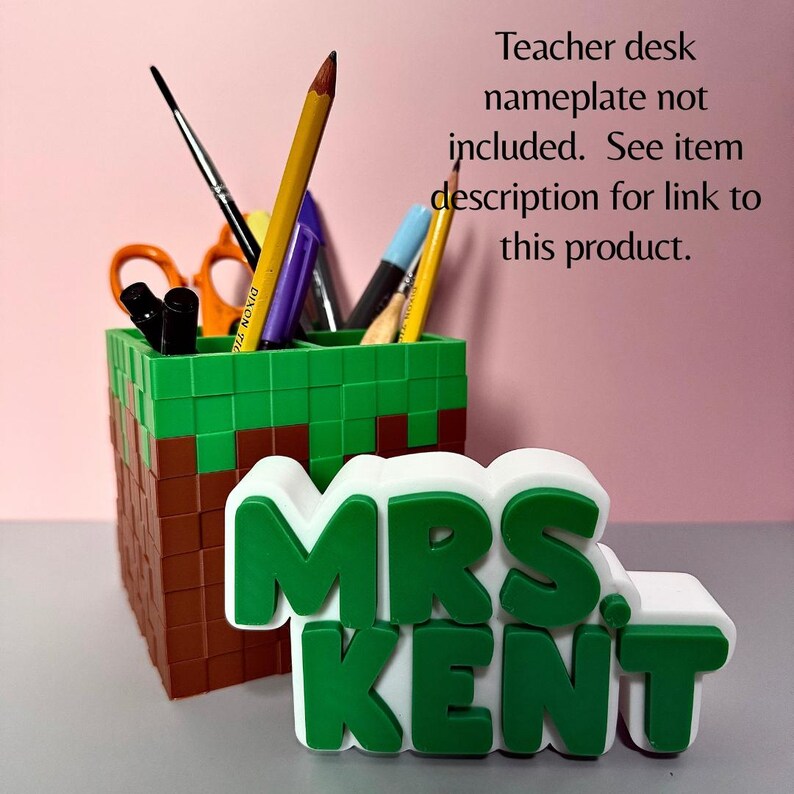 K&ouml;nnte beinhalten: Ein pixeliger Stiftehalter im Block-Design in Gr&uuml;n und Braun, gef&uuml;llt mit verschiedenen Stiften. Davor steht ein wei&szlig;es Namensschild mit erhabenen gr&uuml;nen Buchstaben, die MRS. KENT lesen. Text: Teacher desk nameplate not included.