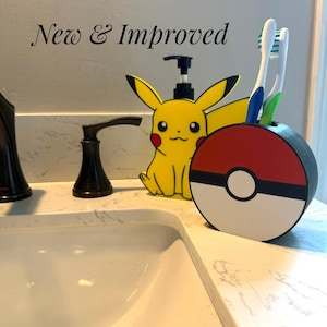 May include: A yellow Pikachu-shaped soap dispenser and a round red, white, and black Poké Ball toothbrush holder containing two toothbrushes. The text New & Improved is visible in the upper left corner above the bathroom sink fixtures.