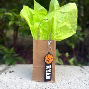 May include: A brown paper gift bag with bright lime green tissue paper and a basketball keychain attached to the handle. The keychain features an orange basketball icon above a black rectangular tag with the name RYAN printed in white letters.