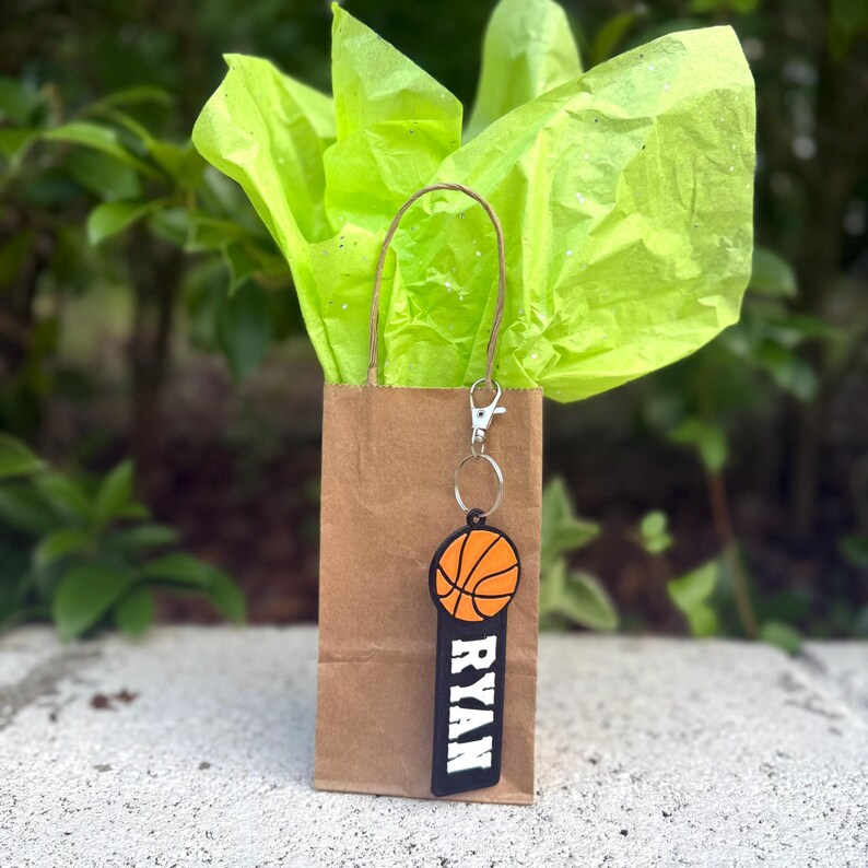 May include: A brown paper gift bag with bright lime green tissue paper and a basketball keychain attached to the handle. The keychain features an orange basketball icon above a black rectangular tag with the name RYAN printed in white letters.