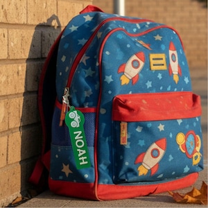 May include: A blue backpack featuring a pattern of white stars and red and white rockets, with a red trim and a front pocket. A green rectangular luggage tag is attached to the zipper, displaying a white truck icon and the name NOAH in white text.